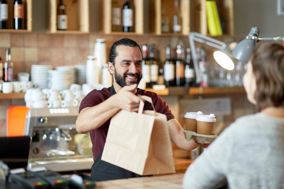A smiling restaurant employee hands two takeout bags across the counter to a customer